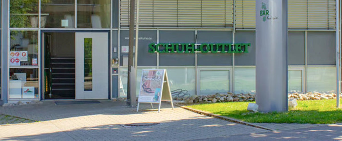 Entrance area of a shoe outlet with large glass windows and a green sign above the door.