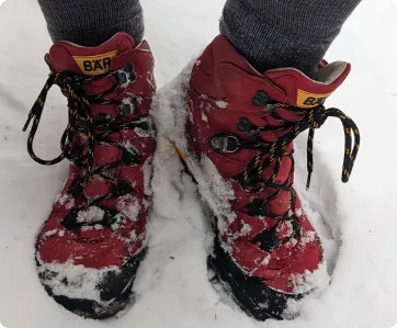 Red hiking boots covered in snow, worn by a person with gray socks on snowy ground.