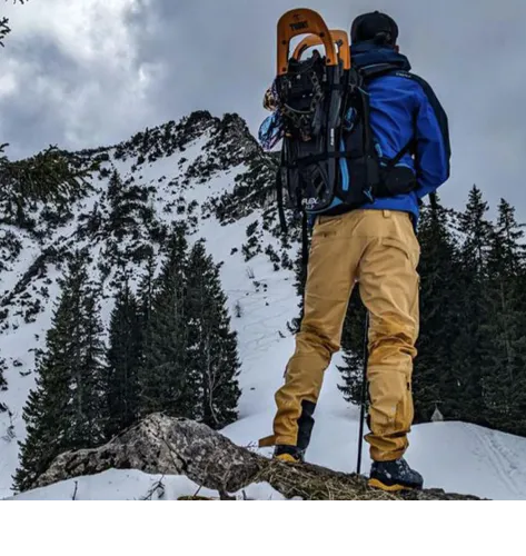 Man in blue jacket and yellow pants with snowshoes in backpack against snowy mountain backdrop
