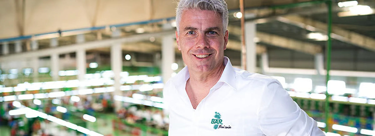 Homme souriant dans un grand bâtiment industriel, portant une chemise blanche avec logo d'entreprise