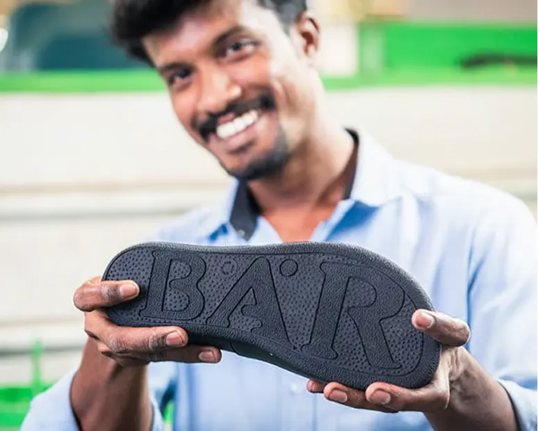 Un homme en chemise bleue tient une semelle de chaussure avec l'inscription BAR face à la caméra
