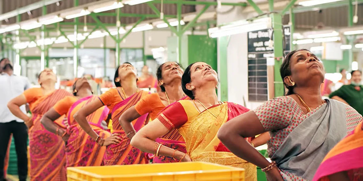 Plusieurs femmes en saris colorés font des étirements dans un hall d'usine. Elles sont alignées et étirent le buste et la tête vers l'arrière.
