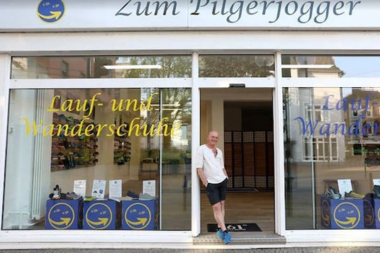 Schaufenster des 'Zum Pilgerjogger' mit Lauf- und Wanderschuhen, Mann steht in der Tür