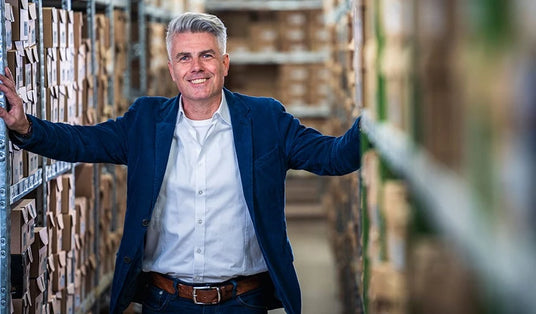 Un homme souriant en jean et blazer se tient dans une allée d'entrepôt entre des étagères remplies de cartons.
